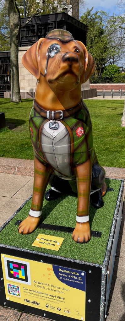 Front view of a sculpture of a guide dog dressed as Sherlock Holmes, wearing a deerstalker hat, monocle, tweed jacket, trousers and boots. The white tag hanging from his brown collar has the address 221B Baker Street on it.