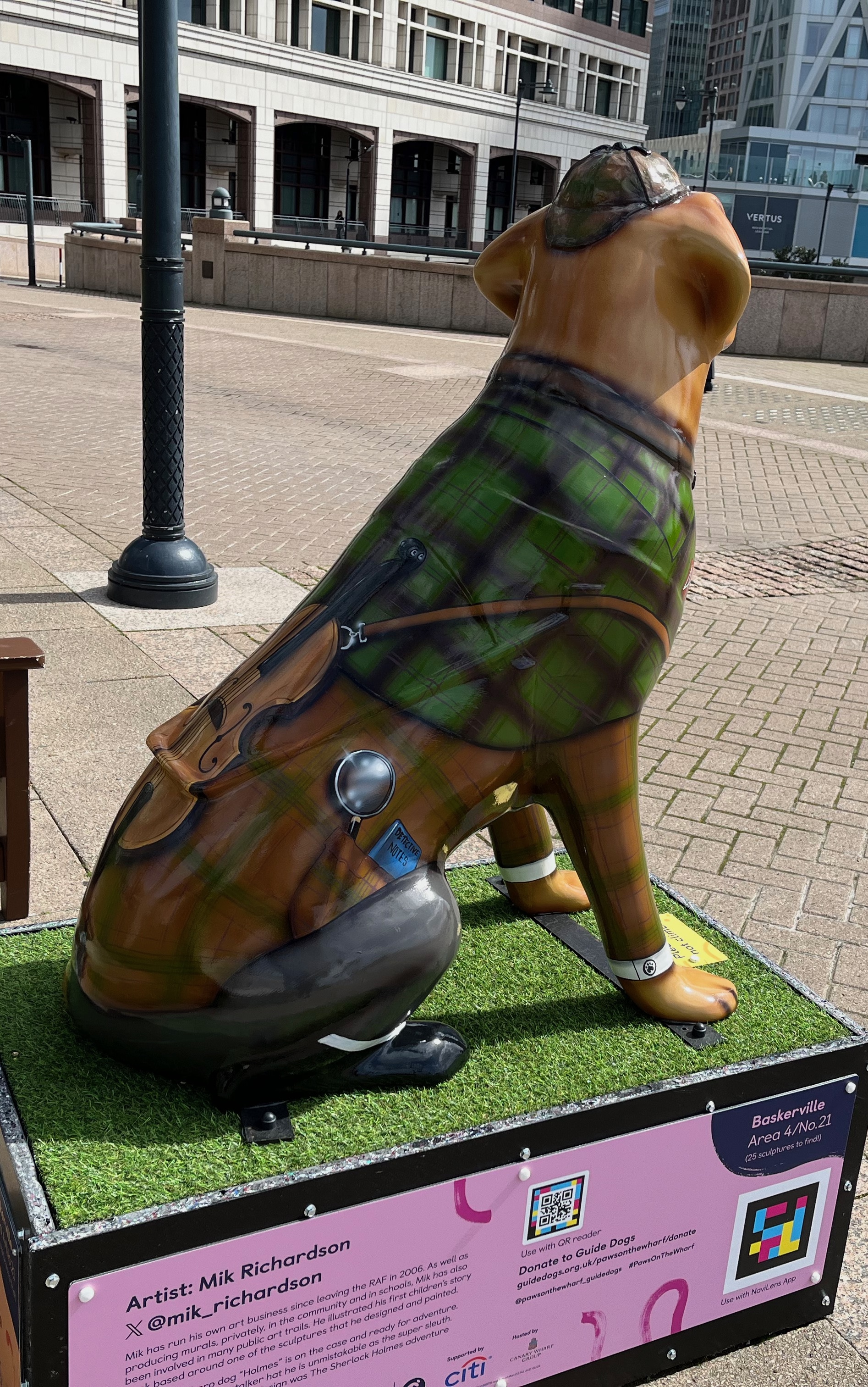 Left view of a sculpture of a guide dog dressed as Sherlock Holmes, wearing a deerstalker hat, monocle, tweed jacket, trousers and boots. A side pocket on his jacket holds a magnifying glass and blue notebook entitled Detective Notes. A violin is attached to the back of the jacket.