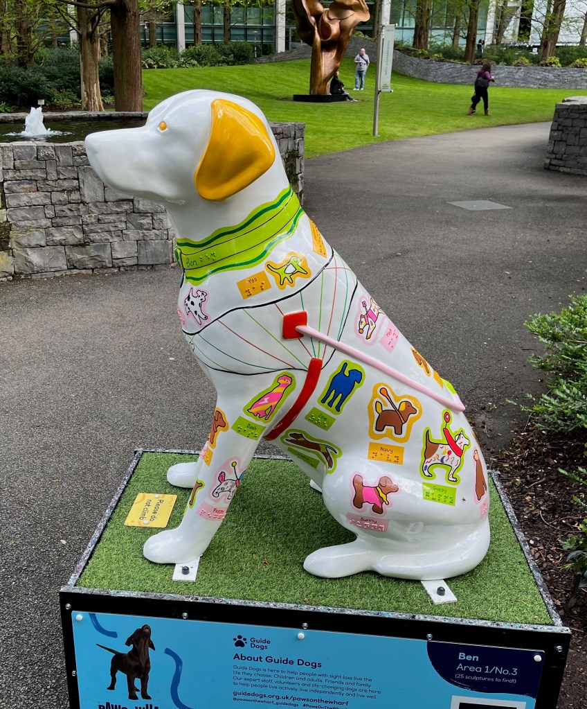 Left side view of a white guide dog sculpture, with a brown ear and green collar. The dog's body and legs are decorated with lots of brightly coloured mini dogs, with their names in Braille underneath, including Yas, Dash, Peggy, Zoe, Coby, Navy, Guppy, Sunny and Jo.