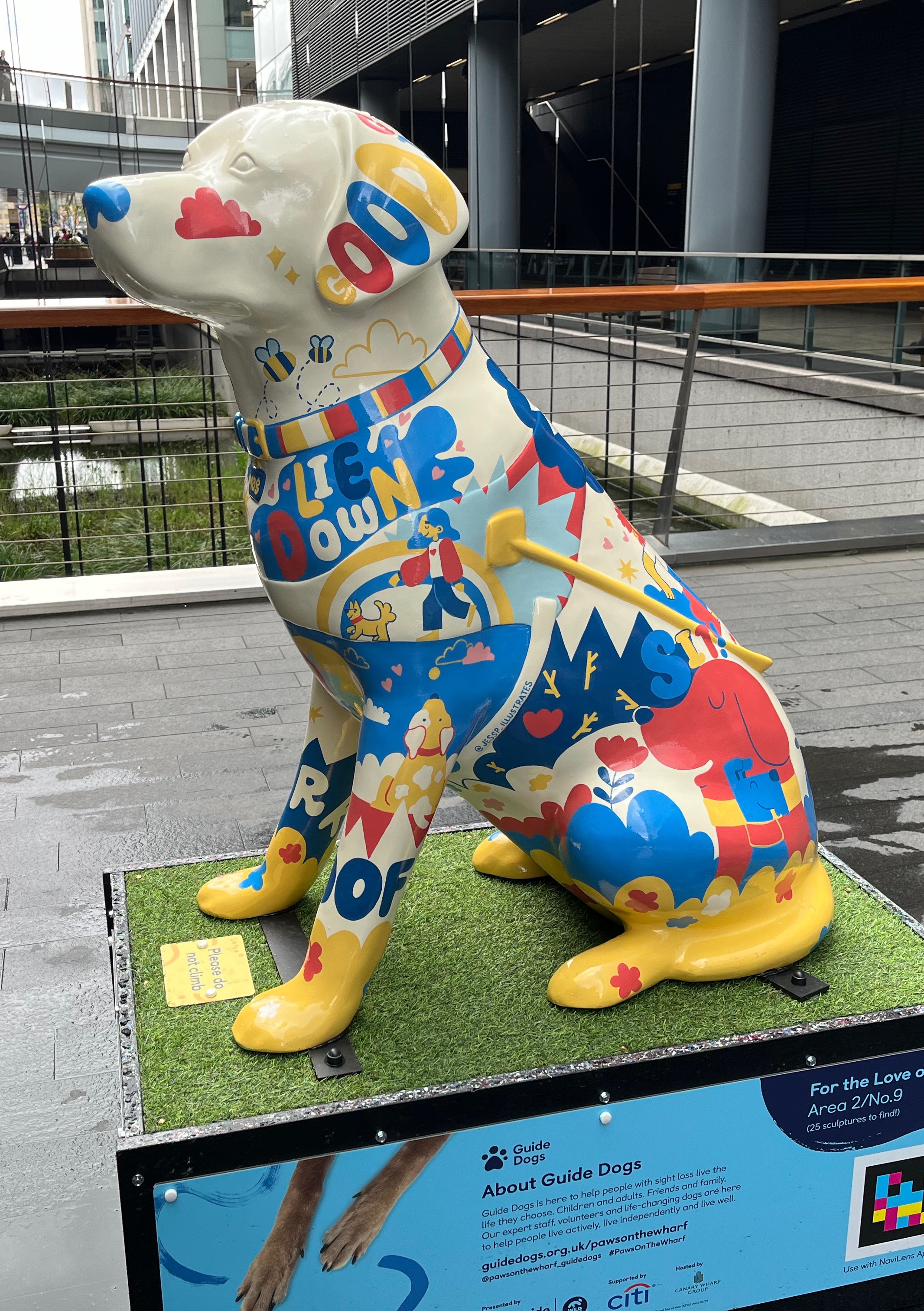 Left side view of a cream coloured sculpture of a guide dog covered in a colourful pattern featuring dogs and their owners, bees, clouds, flowers and hearts, plus phrases including Good, Lie Down, Sit and Woof.