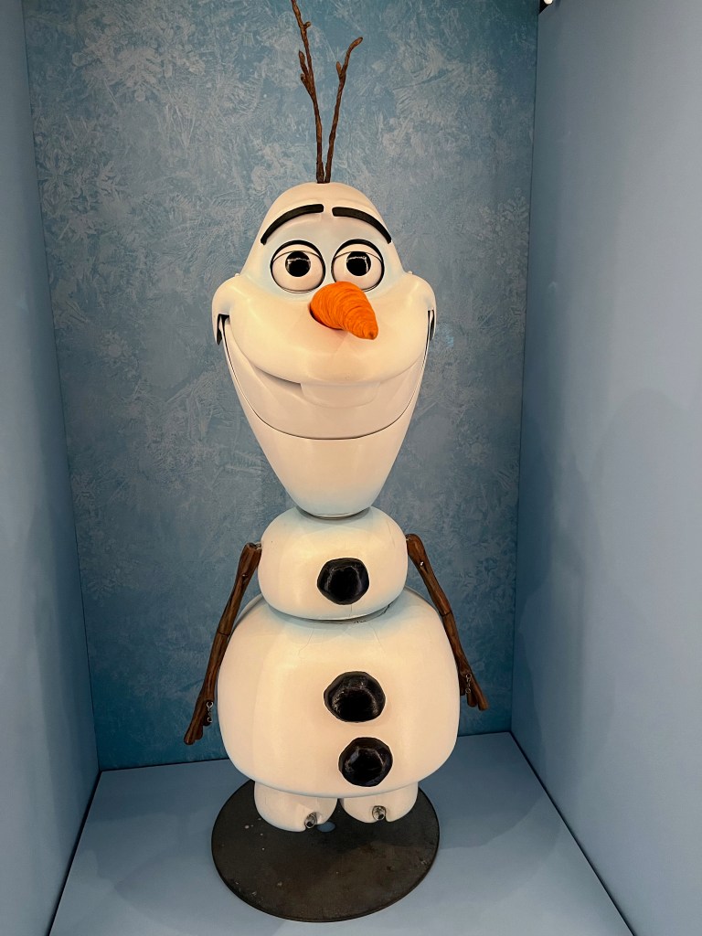 Model of Olaf from the 2013 film Frozen, a large snowman with a tall head and a broad grin on his face, on top of a much smaller ball of snow for the top part of his torso, and then a larger ball of snow for the rest of the torso below that, and finally a small snowball for each foot. He has a carrot for a noise, and 3 big black buttons down his front.