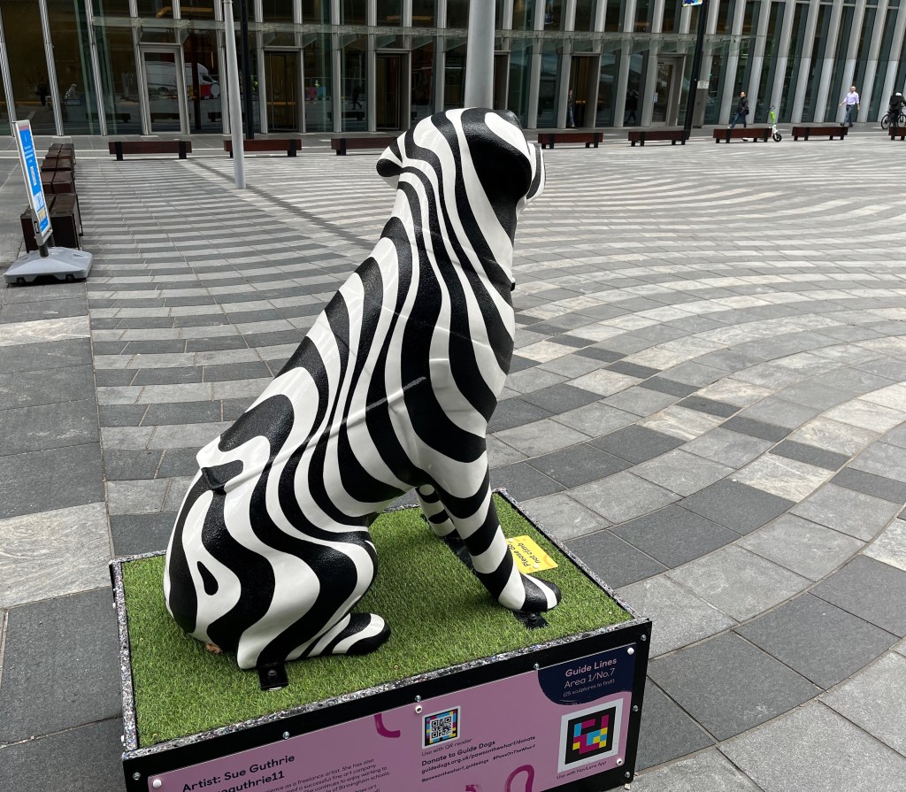 Rear view of a sculpture of a guide dog with a black and white swirly pattern all over it. The black swirls are slightly textured and the white swirls are smooth. The dog is looking out over a large square that has wavy stripes of grey and dark grey paving that complement its own swirling design.
