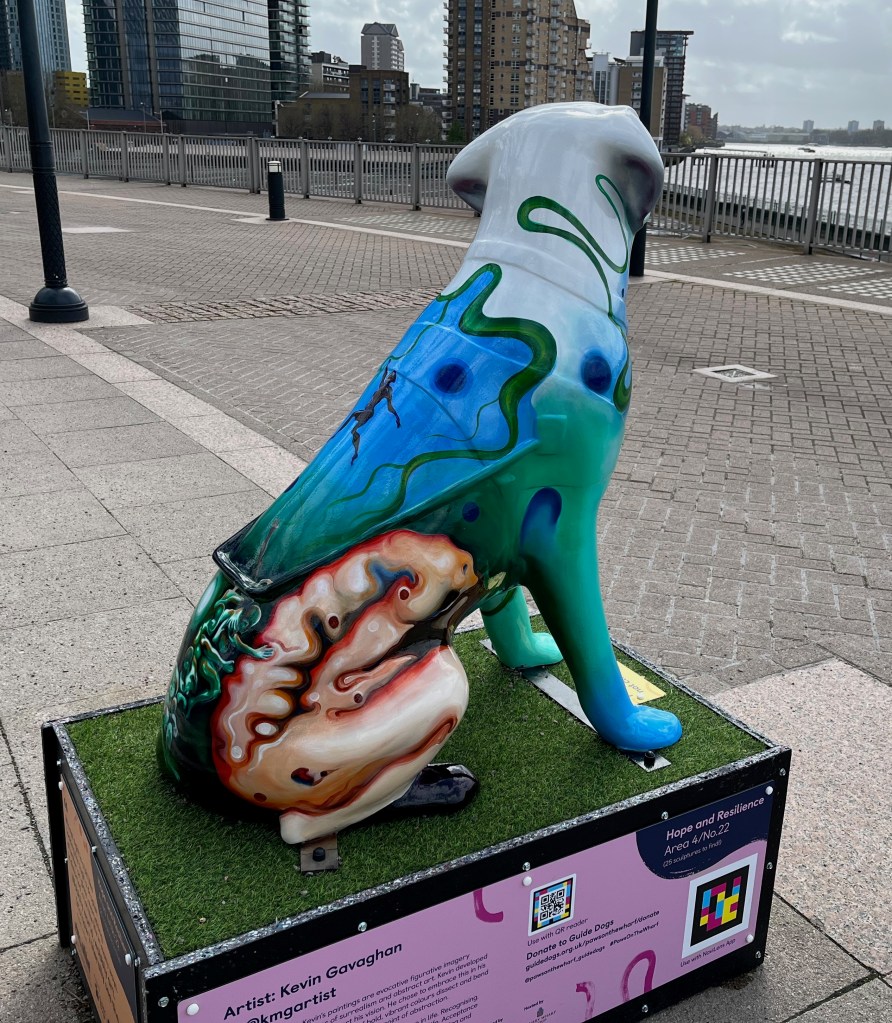 Right side view of a guide dog sculpture with an abstract painting on its hind legs of a person bending over their knees, but being held up by small figures. The dog has blue, green and grey swirls on its upper body.