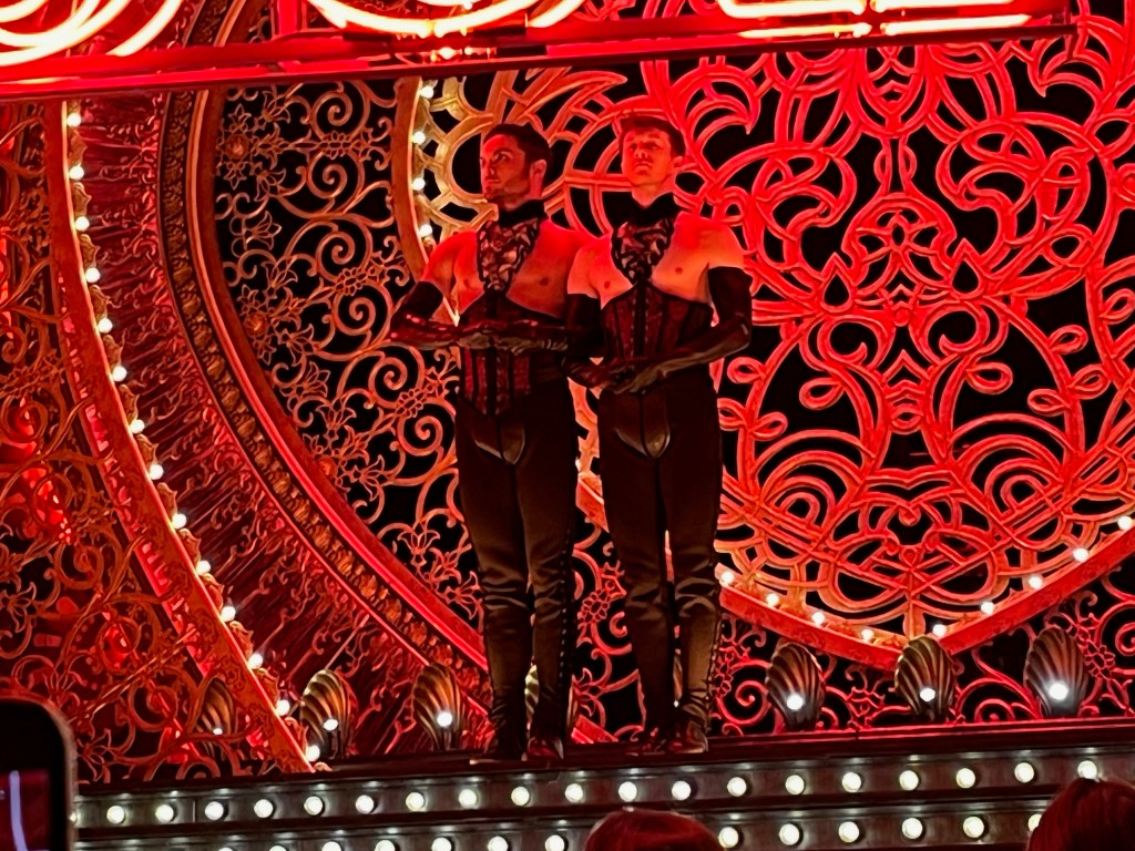 Two men on the stage of Moulin Rouge. Their figure-hugging outfits are entirely black, consisting of trousers, corsets and long gloves that extend to their upper arms. The corsets are connected to their collars by decorative neck pieces, which leave their breasts and shoulders exposed.
