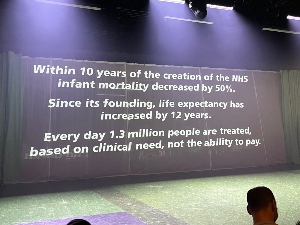 Text projected on to the green hospital curtains at the end of the Nye play at the National Theatre. It says within 10 years of the creation of the NHS, infant mortality decreased by 50%. Since its founding, life expectancy has increased by 12 years. And every day 1.3 million people are treated, based on clinical need, not the ability to pay.