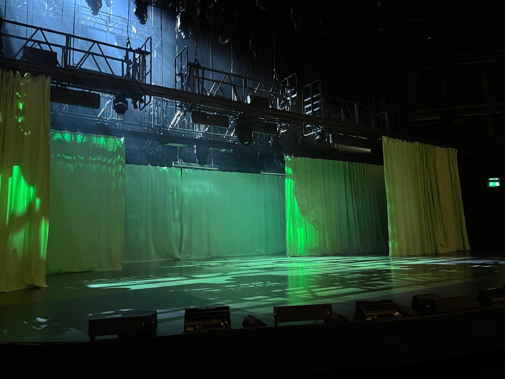 The stage before the start of Nye. A green hospital curtain is fully drawn across the back of the stage, while in front of it a few other curtains are partially drawn across on the left and right sides.