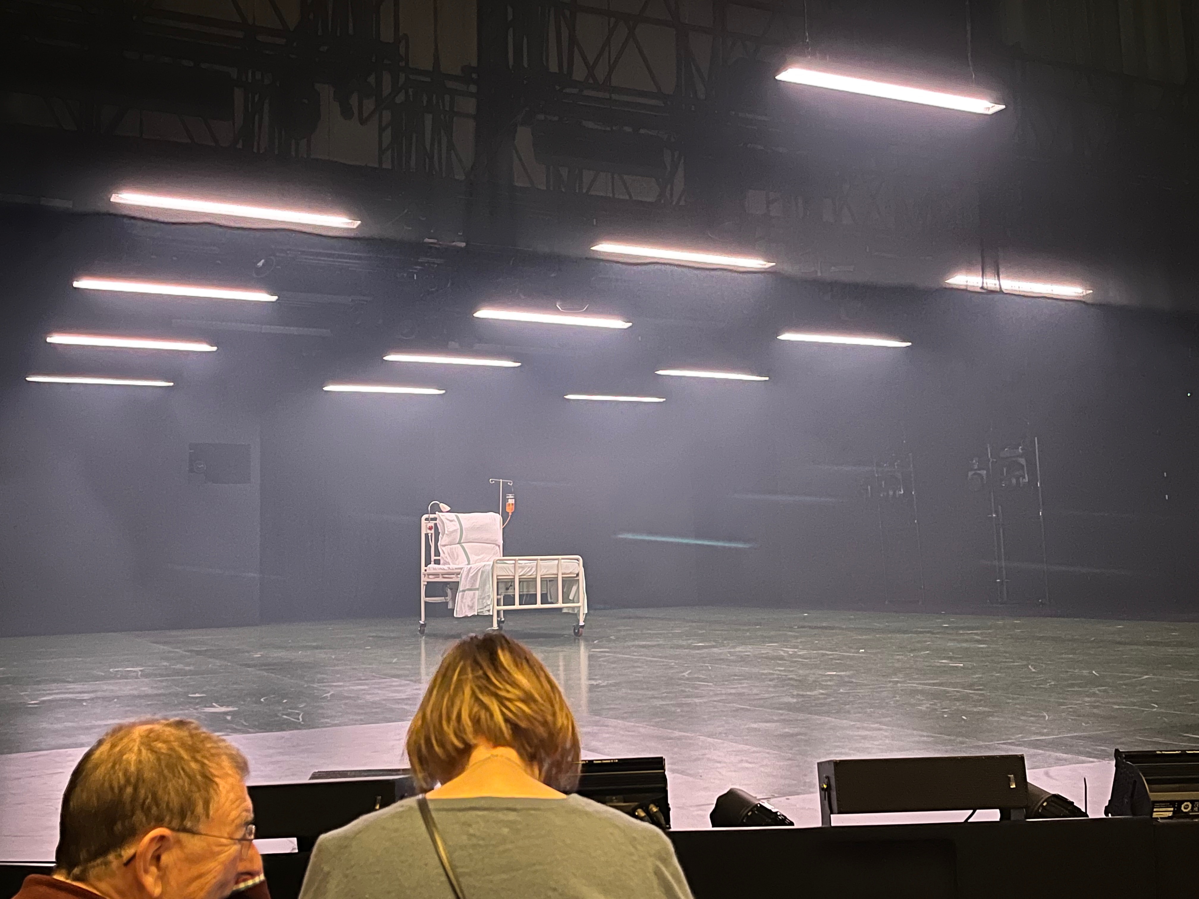 The stage during the interval of Nye, with lights on the ceiling illuminating a space below that is bare apart from an empty hospital bed in the centre of the stage. There is a small lamp attached to one corner of the bedstead, while a bag containing liquid and with a drip tube emerging from it hangs from a metal pole standing next to the other side of the bed.