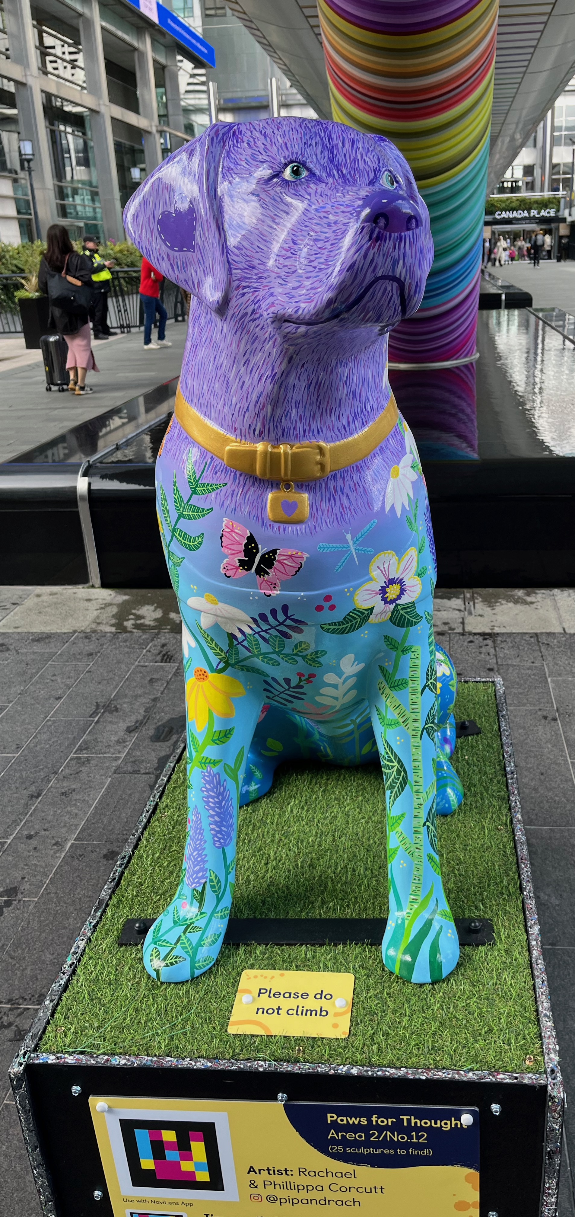 Front view of the sculpture of a guide dog with a purple head fading to a blue body. The body is covered in colourful flora and fauna. The dog is wearing a gold collar with a gold tag attached that features a small blue heart.