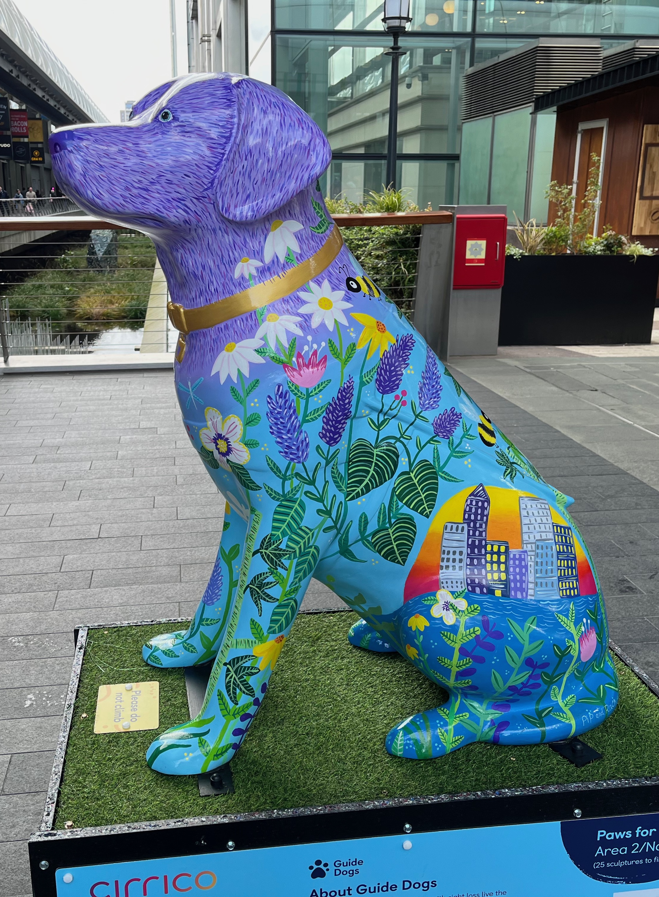 Left side view of the sculpture of a guide dog wearing a gold collar, with a purple head fading to a blue body. The body is covered in colourful flora and fauna and features Canary Wharf cityscape illustrations.