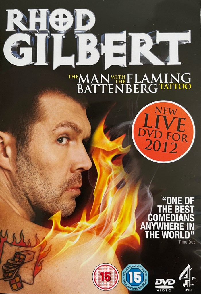 Front cover of the DVD for Rhod Gilbert - The Man With The Flaming Battenberg Tattoo. It shows Rhod glancing backwards as a tattoo on the back of his shoulder of a sliced Battenberg cake on fire catches alight for real with big flames.