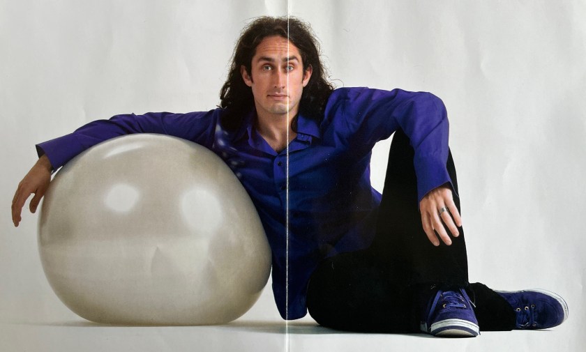 Double-page spread from the booklet with Ross Noble's Fizzy Logic DVD, showing Ross sitting on the floor with his right arm resting on a large inflatable silver ball as he rests against it.