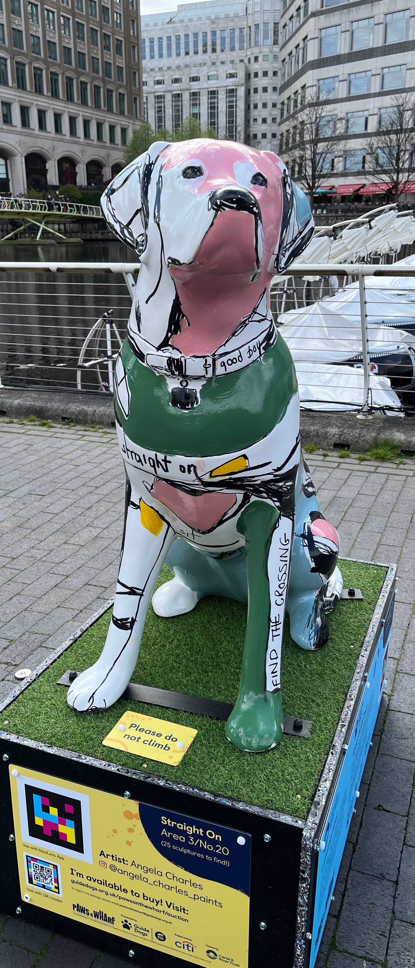 Front view of a sculpture of a white guide dog covered in large patches of green, grey and pink, along with some black markings. The words Straight On are written across its harness, Good Boy on its collar, and Find The Crossing on its left leg.