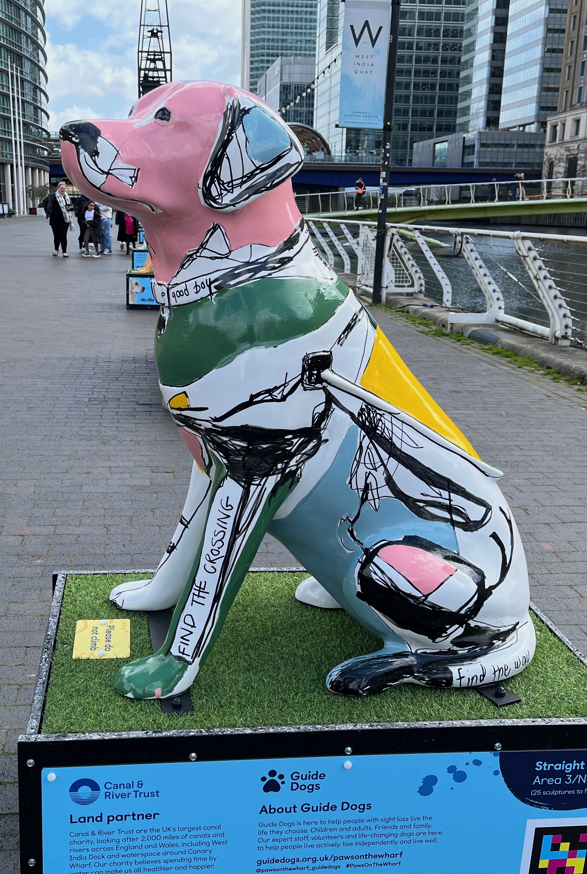 Left side view of a sculpture of a white guide dog covered in large patches of green, grey and pink, along with some black markings. The words Good Boy are written on its collar, Find The Crossing on its left leg and Find The Way on its tail.