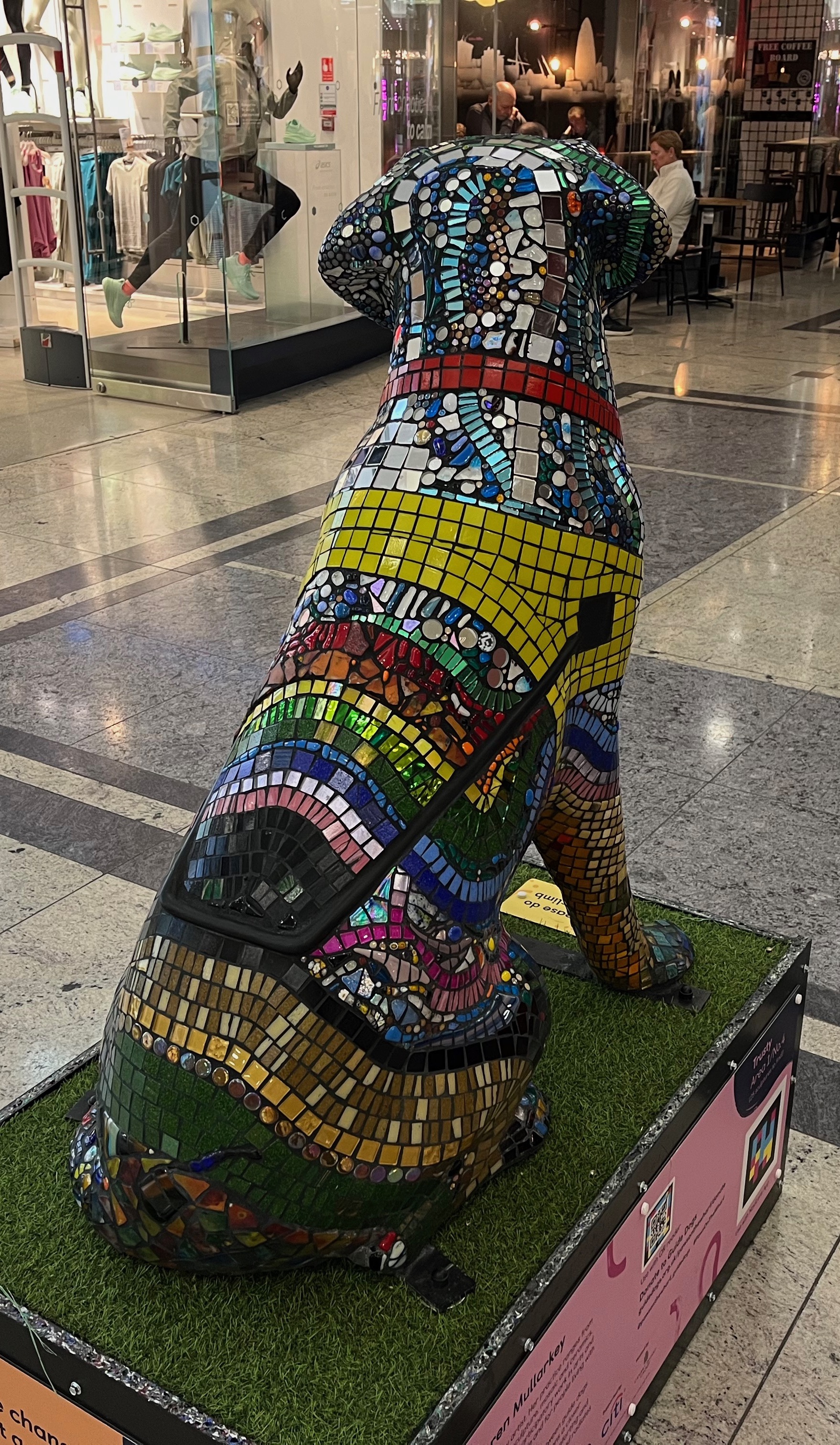 Rear view of a guide dog sculpture covered in small multicoloured tiles making a mosaic pattern, including a yellow mosaic harness with a black handle attached.