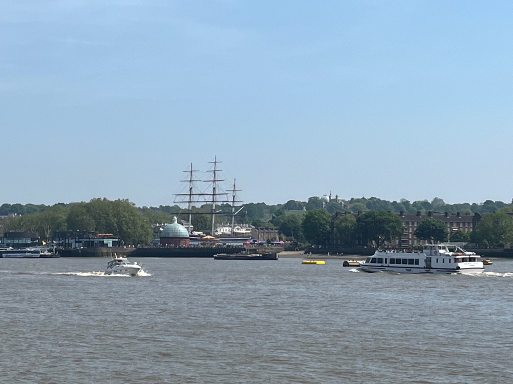 View across the Thames to the Cutty Sark clipper ship with its 3 tall masts, which tower over the round domed building that serves as the entrance to the Greenwich Foot Tunnel.
