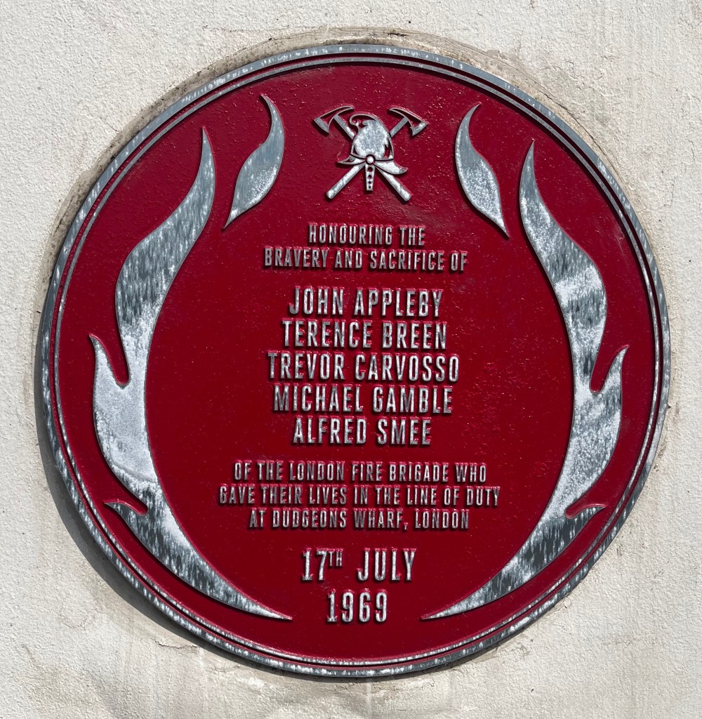 Round red plaque on the Dudgeon’s Wharf Firefighters Memorial. Silver flame motifs run up the left and right side, while in the centre the text says Honouring the bravery and sacrifice of John Appleby, Terence Breen, Trevor Carvosso, Michael Gamble and Alfred Smee of the London Fire Brigade, who gave their lives in the line of duty at Dudgeon's Wharf, London, 17th July 1969.