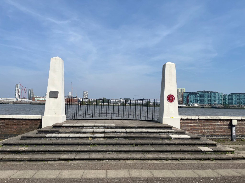 The Dudgeon’s Wharf Firefighters Memorial. 6 steps, gradually decreasing in width, lead to a paved balcony with a curved railing overlooking the Thames. On the left and right of this balcony are two tall white columns, one with a square metal plaque, the other with a round red plaque.