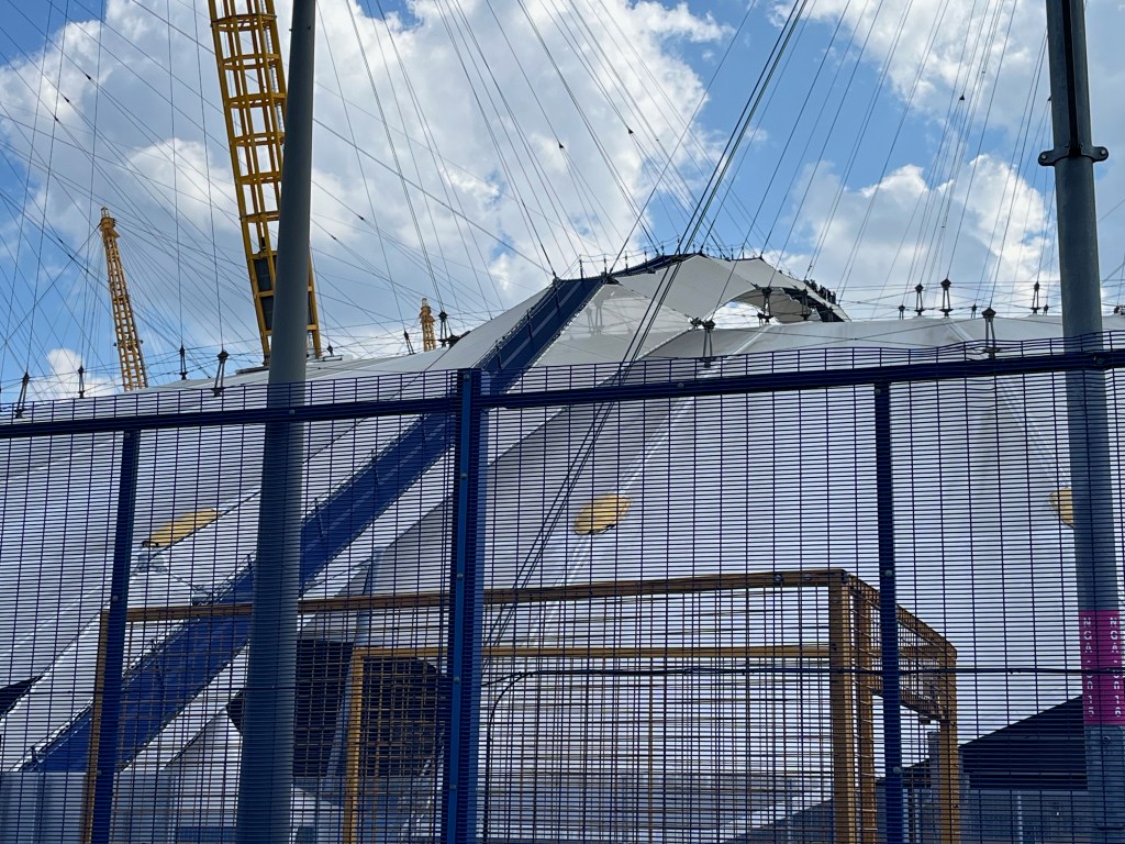 A narrow dark blue pathway with railings that goes up to the roof of the O2 Arena. A group of visitors can be seen at the very top in the distance, having climbed the path to admire the views.