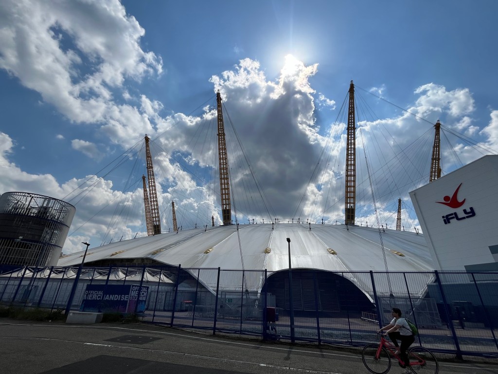 The O2 Arena, with the sun reflecting off its large white dome structure that has tall girder-like structures sticking up high from its roof, that have many cables coming off them attached to the roof. To the right of the arena is the iFly building for indoor skydiving.
