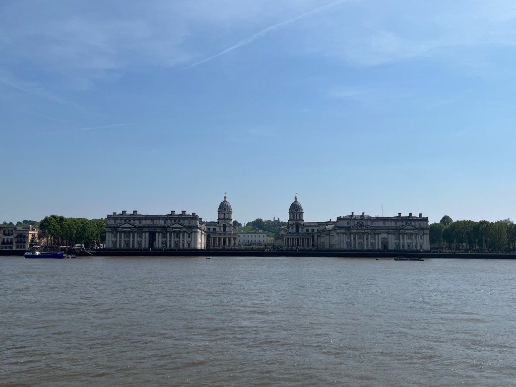 The buildings of the Old Royal Naval College, with two domed towers in the central section, viewed from the other side of the Thames.
