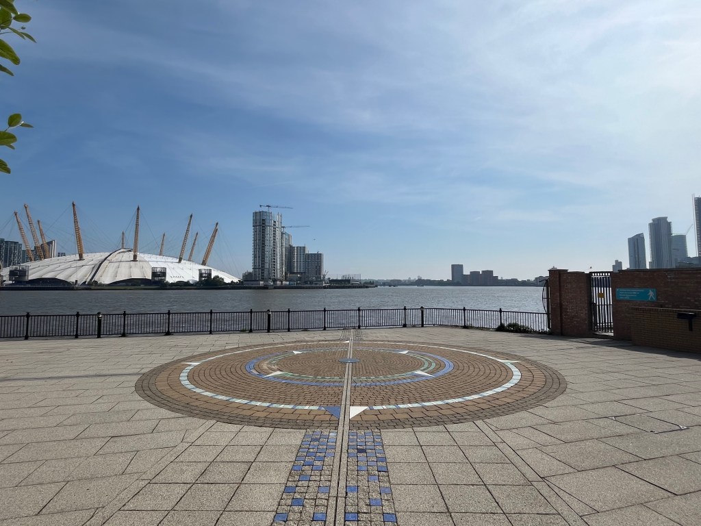 The Prime Meridian Line passing through a large round compass painted on the paving, next to some railings overlooking the Thames. The huge white dome that is the O2 Arena is to the left on the other side of the river.