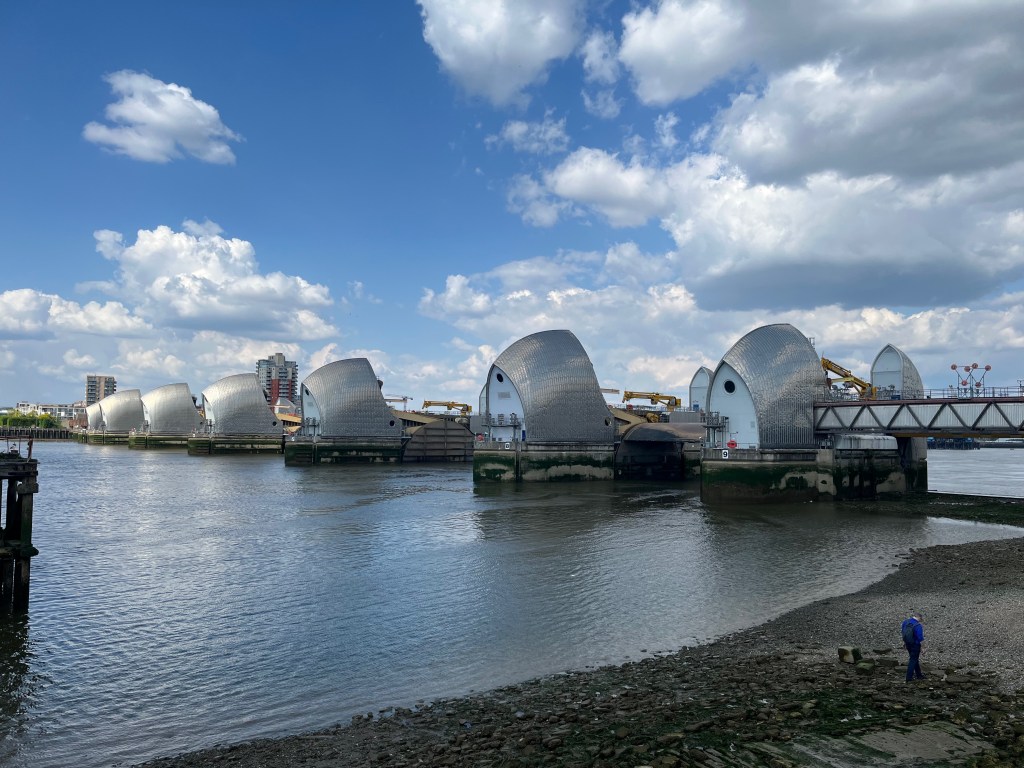 The Thames barrier viewed from the west side, consisting of 9 piers with gaps between them for boats to pass through. Each pier has a domed metallic structure, with a white panel facing outwards that has a round porthole-like window on it. Each pier has the number 1 to 9 on its lower wall, with 9 nearest to us on the south bank.