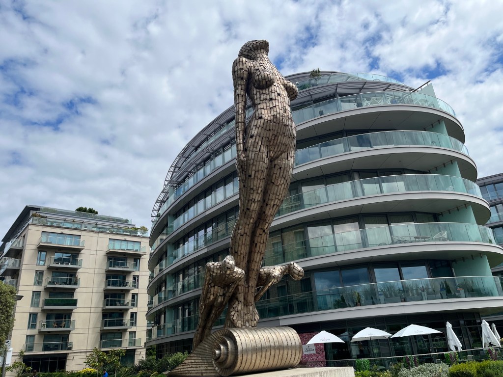 Figurehead statue by John Kirby, viewed from an angle at the front right of the statue. Symbolising the figurehead of a ship, a large female figure leans forward, while two smaller figures are at her feet standing on a shell-like base.
