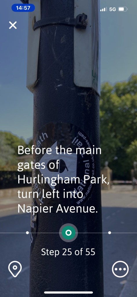 Screenshot of the Go Jauntly app. An image of a large black pole, with large white arrow pointing in the direction of the Thames Path, is overlaid by text that says before the main gates to Hurlingham Park, turn left into Napier Avenue.