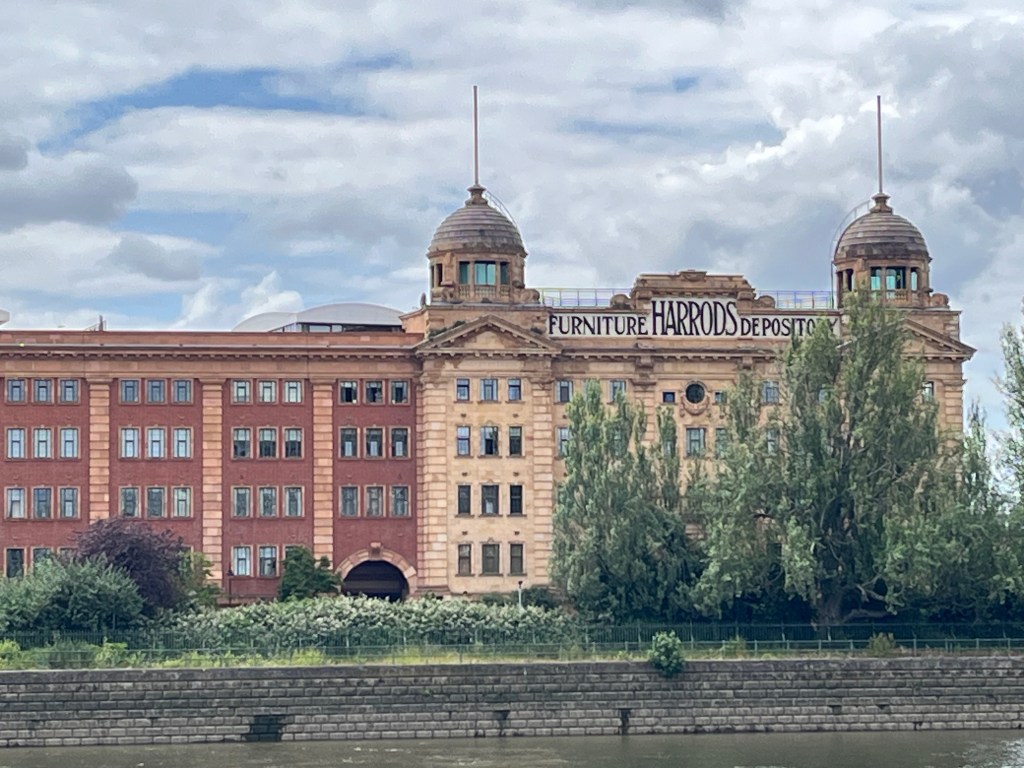 Close-up of one end of the historic Harrods Furniture Depository, a long 4 story building by the Thames. There are two rooms under domes on the roof, either side of a sign along the edge of the roof that says Harrods Furniture Depository in black text on a white background. The word Harrods is enlarged in the centre, in between the other two words in slightly smaller lettering.