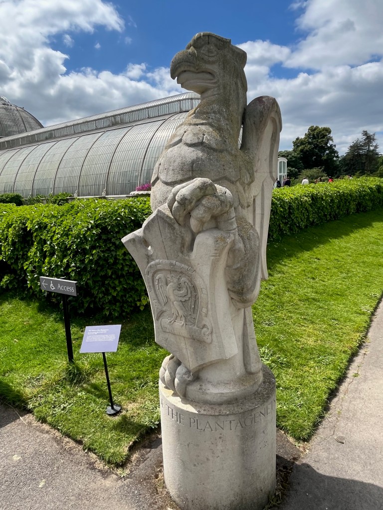 A large stone statue of a falcon standing tall, holding a shield in front of its body. it's called The Falcon Of The Plantagenets by James Woodford.