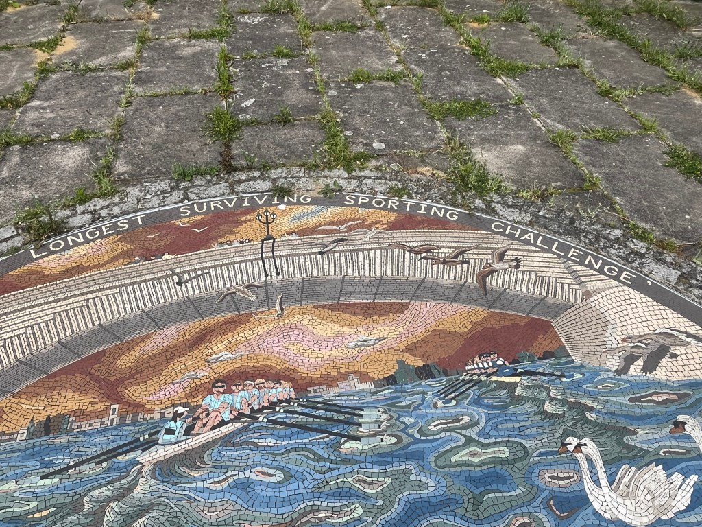 Close-up of the top of the Oxford and Cambridge Boat Race mural, showing small figures of people on a bridge cheering on the two teams of rowers passing beneath them, while birds fly by.