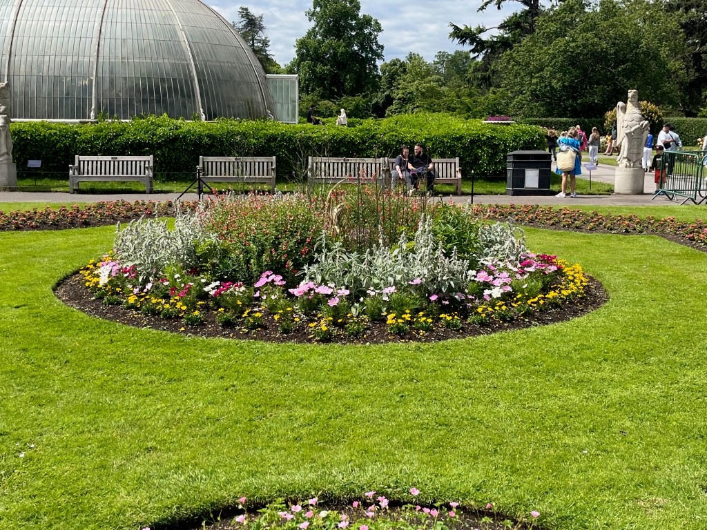 A round flowerbed with greenery surrounded by pink, white and yellow flowers.