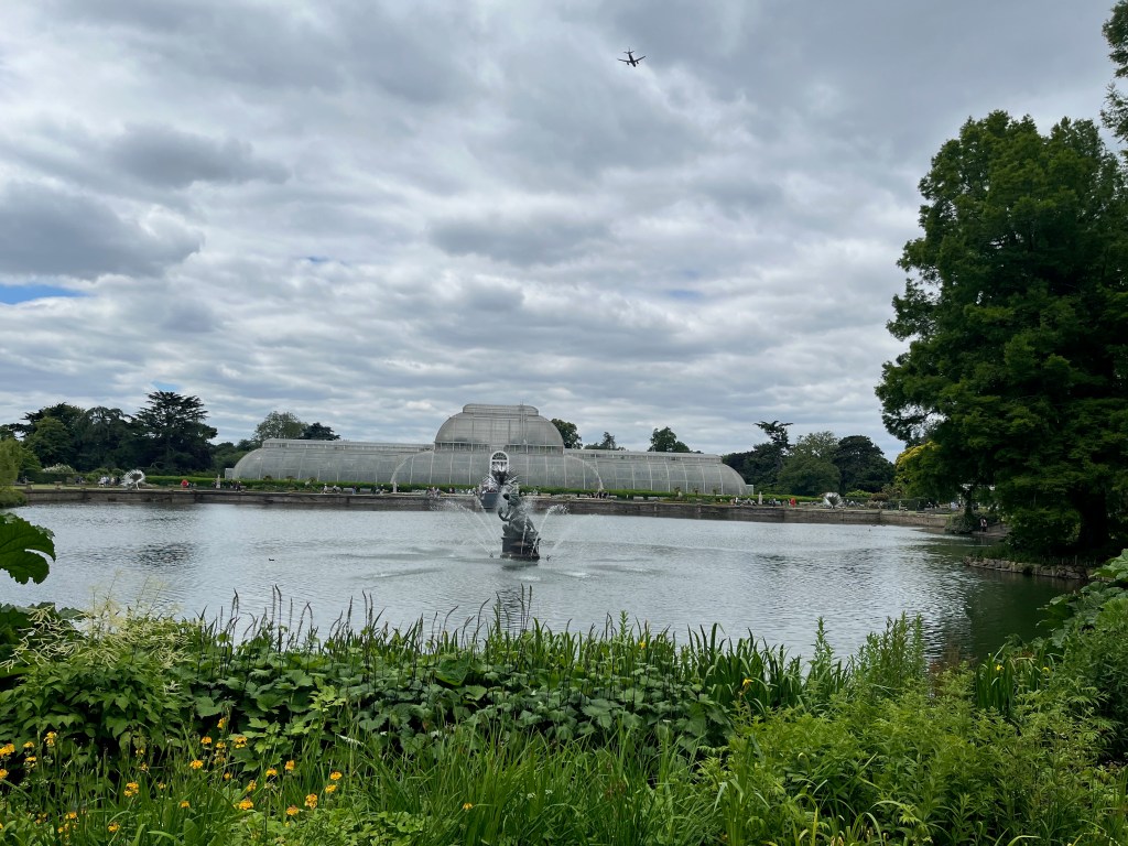 A view across a large pond in Kew Gardens to Palm House, a large glasshouse, in the distance. A fountain in the middle of the water has curving jets of water spraying out of it.