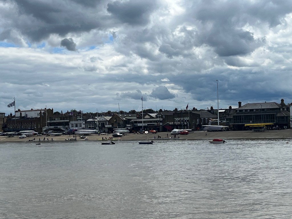 A view across the River Thames to a wide strip of land with rowing boats and boating school buildings.