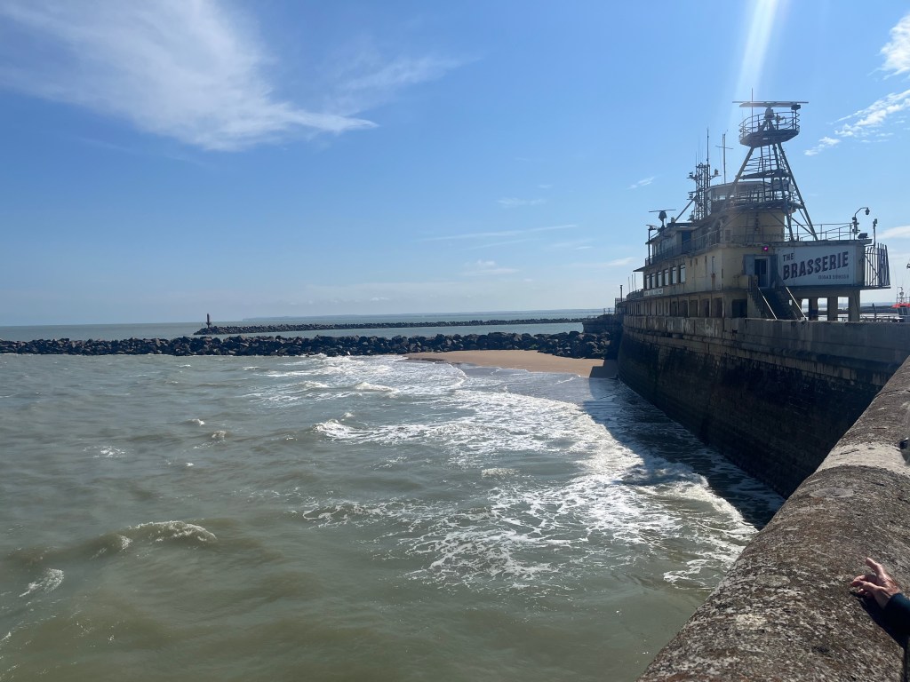 Brasserie building on an outer arm of Ramsgate Harbour. The walkway leading up to it, and a small wall stretching off from the opposite corner of the structure, form a corner in which waves are lapping at a tiny beach.