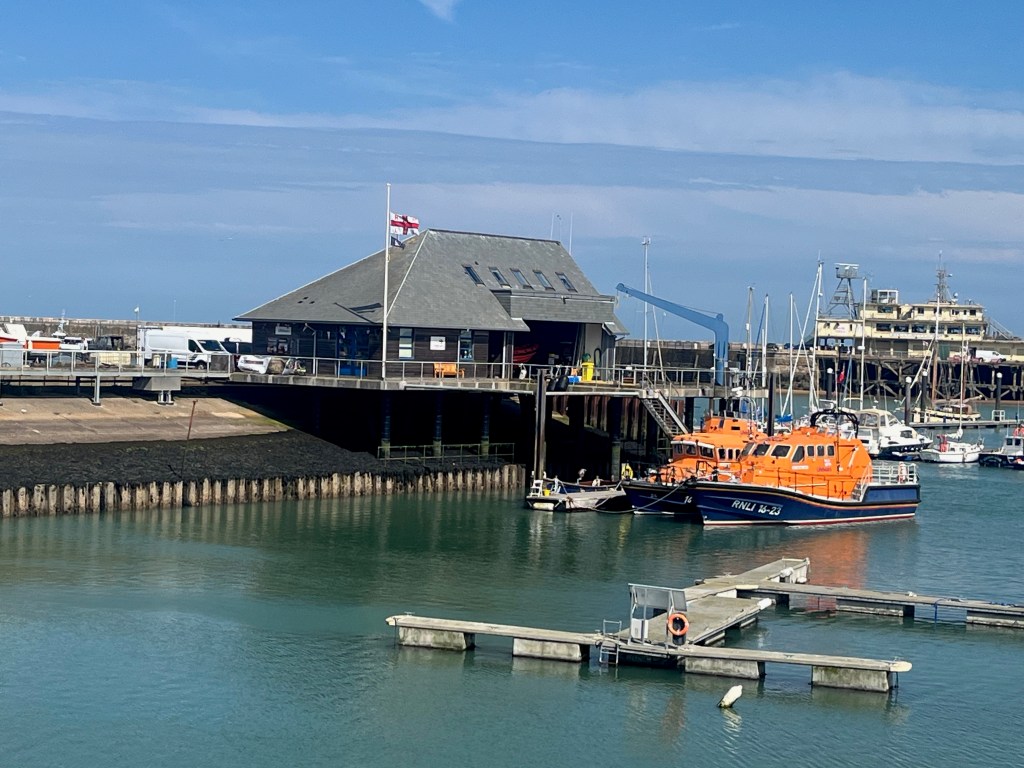 RNLI lifeboat station in Ramsgate Royal Harbour, with one of their boats parked next to it, painted mainly orange but with a dark blue underside.