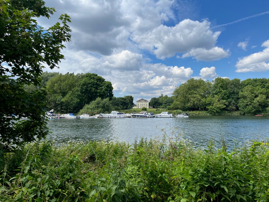 View across the River Thames to Marble Hill, a white three-storey house with a triangular roof. It sits on an area of grass in a clearing between large groups of trees. Lots of boats are moored along the opposite bank as well.