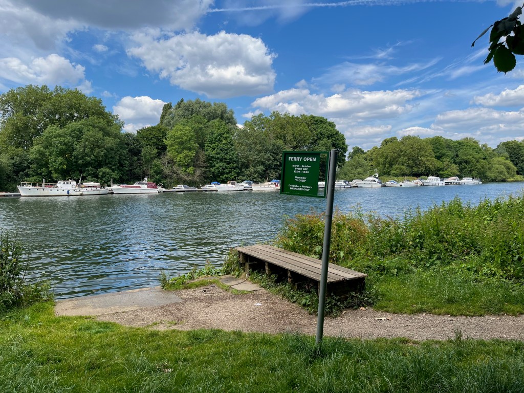 A small slope leading down to the edge of the River Thames, with a bench that for people to sit on. A green sign attached to a pole has white text that reads Ferry Open. March to October, every day 10am to 6pm. November closed. December to February, weekends only.