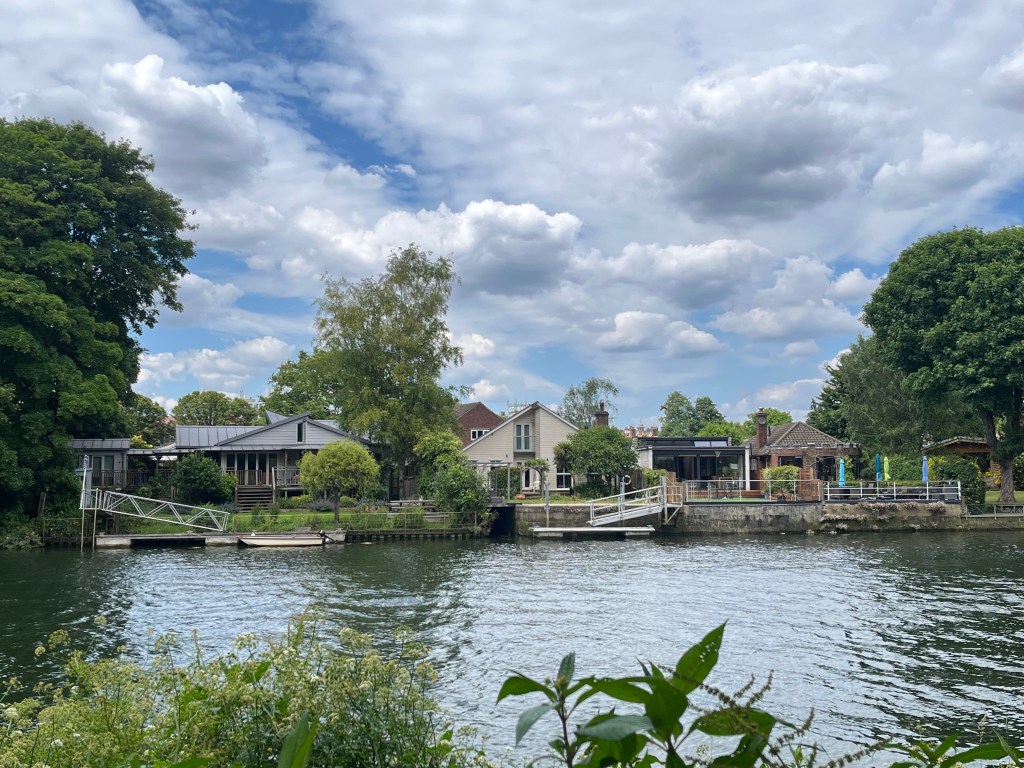 A few bungalows and a two-storey house on the other side of the River Thames, with a couple of sloping walkways leading down to small jetties, one of which has a small boat moored next to it.
