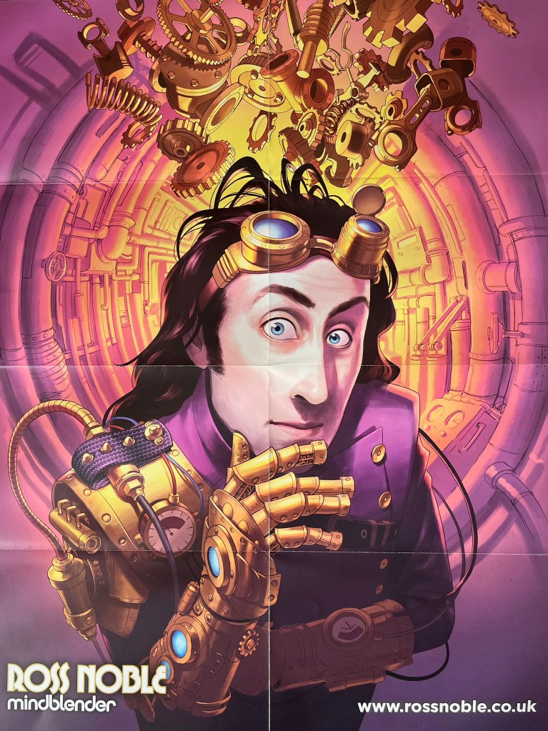 Poster from Ross Noble's Mindblender DVD. It shows an image of Ross stroking his chin thoughtfully with his hand, but it's a gold robotic arm with nuts and bolts, a pressure dial, and a couple of blue lights. His other hand held across his chest in shadow, is similar. He's wearing a purple jacket and has a pair of gold goggles pulled up on to his forehead. Behind him is a circular corridor lined with lots of pipes, bathed in a purple light around Ross but becoming yellow in the distance. At the top of the corridor, are wheels and cogs and other machine elements.