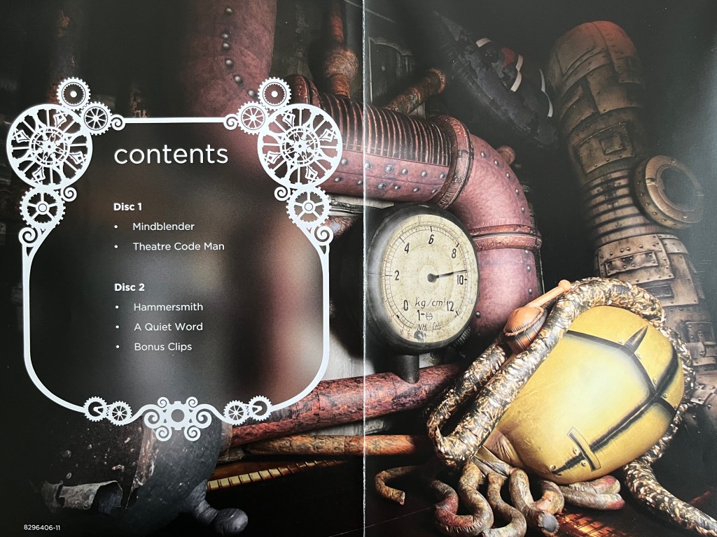 Double-page spread from the booklet with Ross Noble's Mindblender DVD, with a picture of the stage set that looks like a strange factory, with pipes, a large dial and other machinery. On top of this, on the left page, is a list of the contents of the 2 discs in the set. Disc 1 has Mindblender and Theatre Code Man, while Disc 2 has Hammersmith, A Quiet Word and Bonus Clips.