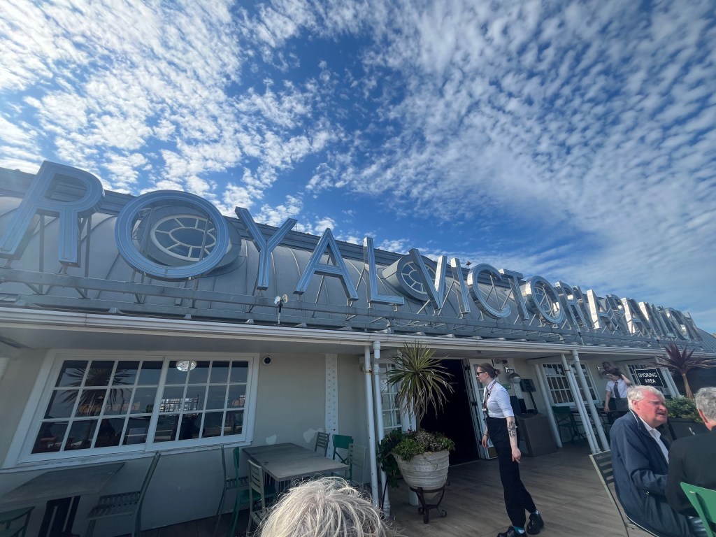 The outside terrace on the upper floor of the Royal Victoria Pavilion pub in Ramsgate. Large metal letters spelling out Royal Victoria Pavilion stand in front of the roof, with the sunlight reflecting off them.