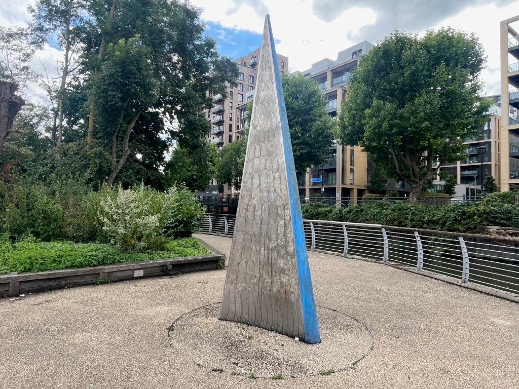 Artwork called Sail by Sophie Horton. It is a large sculpture in the shape of a dinghy sail. The silver surface facing us is convex and resembles the scales and tail of a fish.