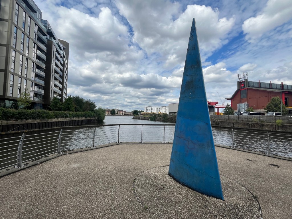 Artwork called Sail by Sophie Horton. It is a large sculpture in the shape of a dinghy sail. The blue side facing us has a convex surface and the design of a sail's seams. Beyond the sculpture, a wide waterway leads out into the River Thames.