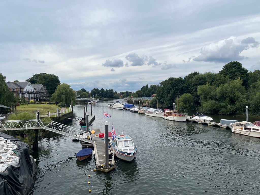 View of the Thames from the Teddington Lock footbridge. In front of the bank nearest us, a white boat is moored against a small jetty, which is connected to the shore by a walkway that slopes up then flattens out, forming a short bridge over the water. A small Union Jack flag is flying on a pole on the jetty. On the far bank, several boats are moored up in a line in front of some large bushy trees.