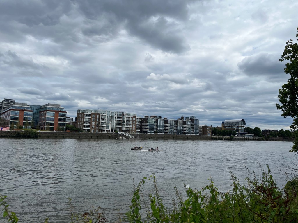A view of the River Thames, with apartments on the opposite bank. Two people can be seen in a rowing boat, followed by a person on a smaller boat who is giving them instructions.