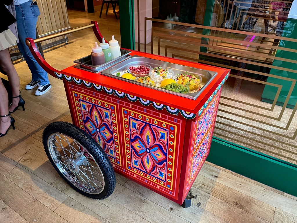 A buffet trolley with 2 big wheels on each side at the back, and a small wheel underneath in the centre at the front. It is very artfully decorated with bright colours surrounded by red framing. On the top of the trolley are dishes of colourful foods, with white, red, green and yellow being the main colours.