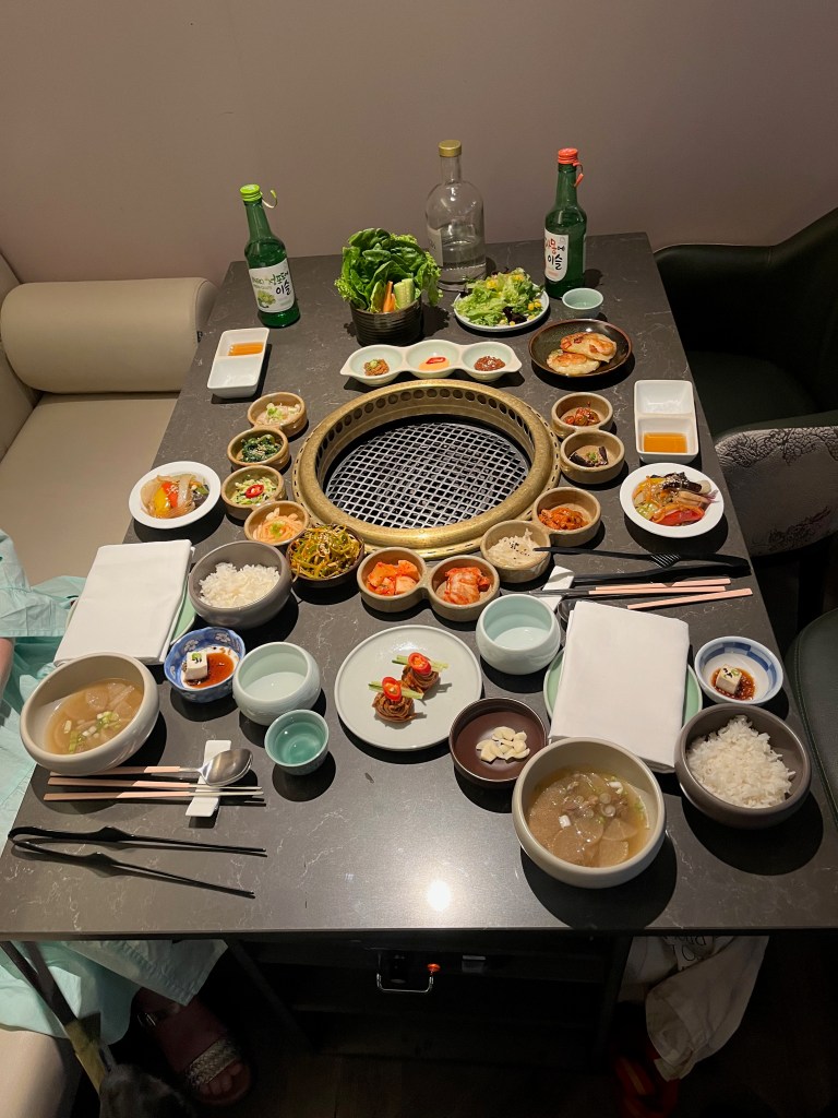 A table full of lots of small bowls and plates with various appetisers and sides at the Korean Grill Kensington restaurant. In the centre of the table is a large round opening with a grate, on which meat will later be grilled.