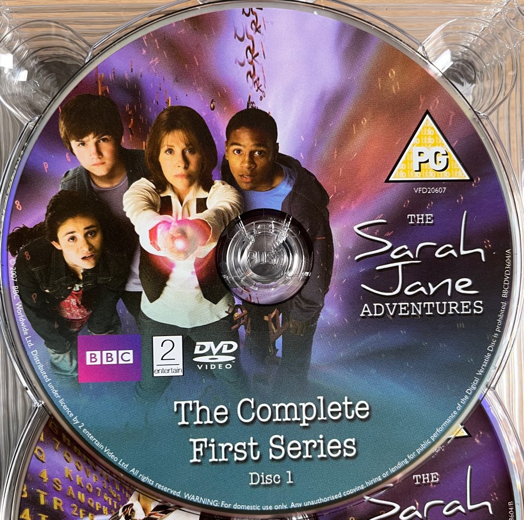 Series 1 Disc 1 from the DVD box set of The Sarah Jane Adventures, including a photo of Sarah as she points her sonic lipstick directly ahead of her, while surrounded by Maria, Luke and Clyde.