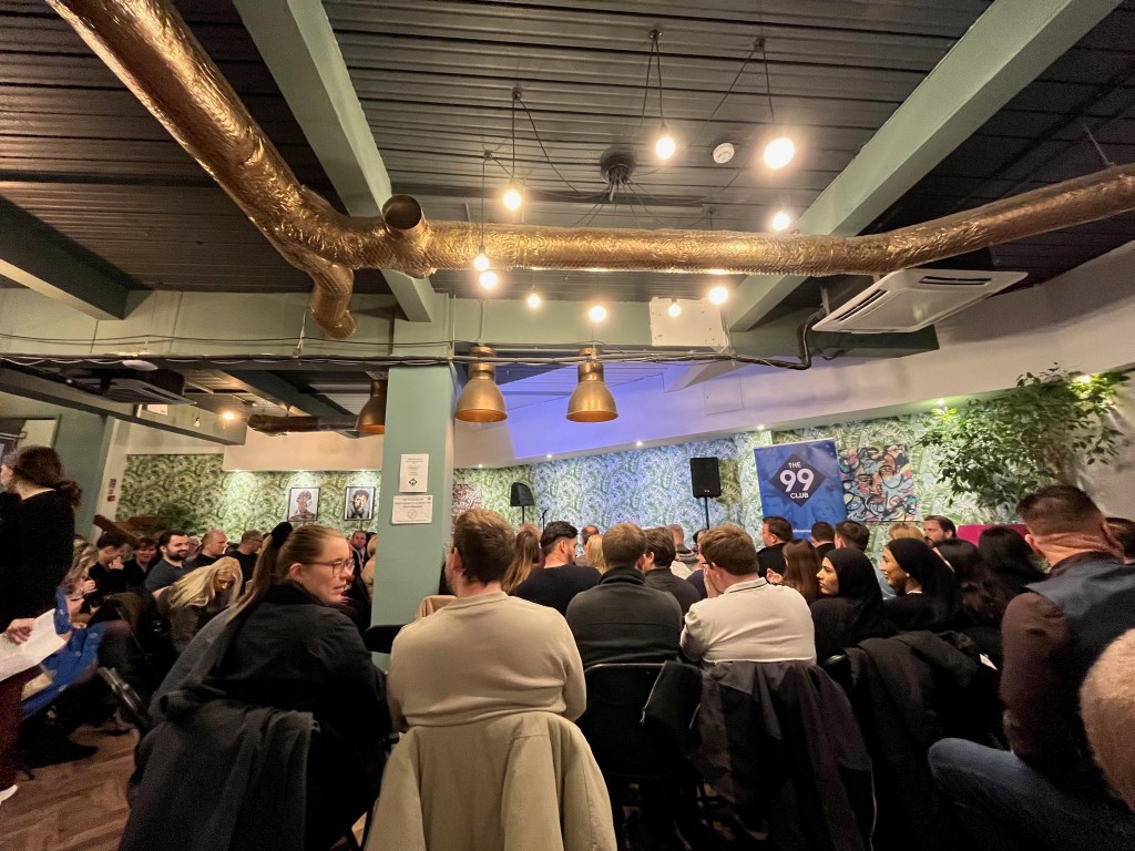 The 99 Comedy Club in the Arboretum Leicester Square. It's a brightly lit basement room, with a large metal pipe going across the ceiling as well as the lighting. The audience are on rows of seats facing the stage on the far wall.